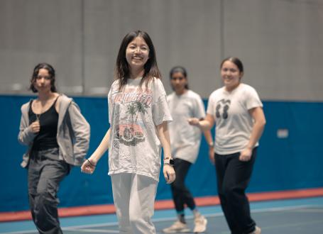 A group of smiling university students with a girl in focus in the centre. She is fair with dark brown hair wearing round silver glasses and a baggy tshirt, smiling big looking to the side.